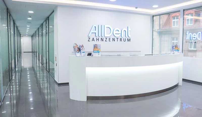 Empfangstresen in modernem Zahnarztzentrum mit beleuchtetem Schriftzug 'AllDent ZAHNZENTRUM' an wei&szlig;er Wand und Glasw&auml;nden im Flur.
