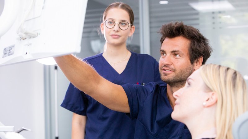 Eine Oralchirurgin und ein Oralchirurg zeigen einer Patientin einen Befund auf einem Röntgenbild